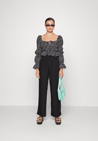 Blusa preta com padrão floral, mangas bufantes e detalhe de laço, combinada com calças largas pretas. Acessórios incluem uma mala verde menta e sandálias de plataforma.