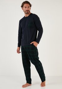 Mørkeblå langærmet top med en grønternet lomme, parret med grønternede pyjamasbukser, løs pasform og af bomuldsstof, barfodet på en lys baggrund.