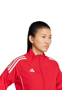 Rote Adidas-Sportjacke mit weißem Drei-Streifen-Design, hohem Kragen und einem Reißverschluss vorne. Glatter Stoff und sportliche Passform.