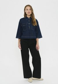 Ženska, ki stoji, oblečena v temno modro denim srajco, široke črne hlače in bež mokasinke, proti enostavnemu belemu ozadju.