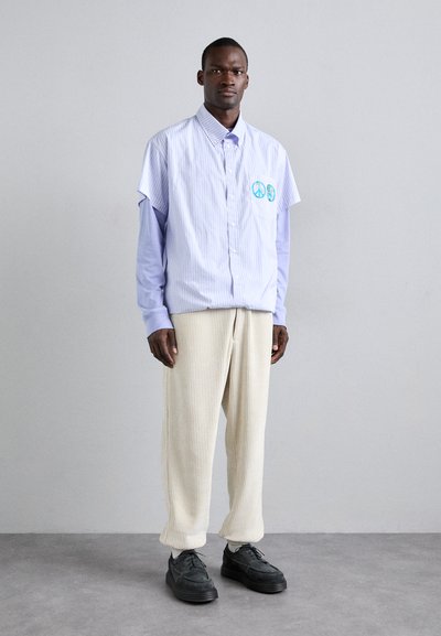 Camicia a maniche corte blu e bianca a righe con simbolo di pace ricamato, sovrapposta a una camicia a maniche lunghe di colore lavanda, abbinata a pantaloni crema a coste e scarpe nere.