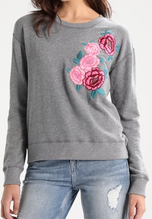 Femme portant un pull gris à manches longues avec une broderie florale rose et rouge sur la poitrine, associée à un jean bleu clair déchiré.