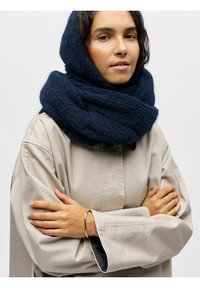 Écharpe en tricot bleu marine foncé enroulée autour du cou, portée avec une chemise oversize beige clair. Présente une texture douce et texturée.