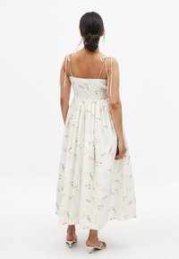 Mujer con cabello oscuro en un moño bajo, vestida con un vestido midi sin mangas de flores blancas y tirantes atados, y sandalias de tacón bajo blancas.