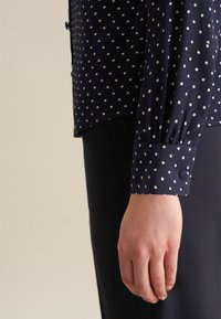 Camicia blu navy con pois bianchi, dotata di polsini con bottoni e una texture liscia. La camicia è abbinata a pantaloni scuri e aderenti.