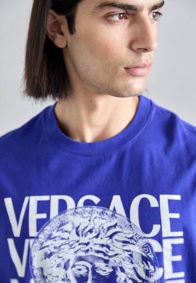 T-shirt en coton bleu avec un col rond ; présente un grand texte blanc "VERSACE" et un graphique de méduse sur le devant. Texture lisse.