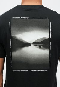 Czarna koszulka z dużym monochromatycznym krajobrazem na plecach, z białym tekstem zawierającym informacje o marce i produkcji.