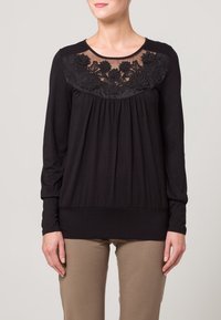 Svart långärmad topp med en genomskinlig, broderad blompanel vid halsringningen och en rynkad framdel med slät tygkvalitet.