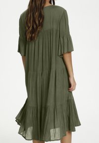 Mujer con cabello largo y ondulado que lleva un vestido midi verde oliva suelto de varias capas con mangas acampanadas, visto desde la espalda contra un fondo simple.