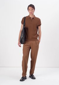 Polo castanho de manga curta com colarinho de zip, combinado com calças castanhas ajustadas. O modelo segura uma mala de tote preta em couro e usa sapatos pretos.