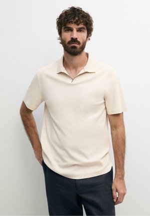 Uomo con capelli ricci e barba in piedi davanti a uno sfondo bianco, indossa una polo beige chiaro a maniche corte e pantaloni scuri.