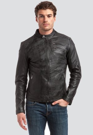 Mann trägt eine schwarze Lederjacke mit Reißverschluss, mit Reißverschlusstaschen auf der Brust und an den Seiten, kombiniert mit blauen Jeans, vor einem grauen Hintergrund stehend.