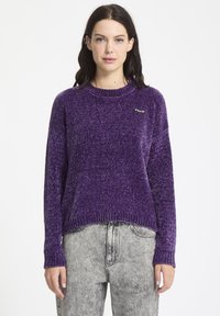 Lilla strikket sweater med en tekstureret overflade, ribbede manchetter og kant, med et lille sort mærke på den øverste venstre side. Sammen med grå jeans.