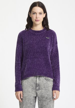 Paars gebreid sweater met een gestructureerde afwerking, ribgebreide manchetten en zoom, voorzien van een klein zwart label aan de bovenkant links. Gecombineerd met grijze jeans.