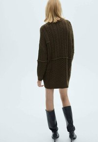 Robe en tricot vert foncé avec des motifs de câbles texturés, manches longues et coupe ample. Associée à des bottes noires jusqu'aux genoux.