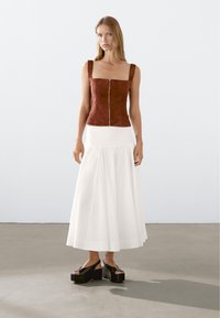 Bruine suède mouwloze top met vierkante halslijn, voorrits, gecombineerd met een witte luchtige rok en zwarte platform schoenen. Soepele texturen.