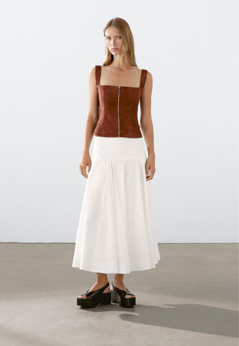 Bruine suède mouwloze top met vierkante halslijn, voorrits, gecombineerd met een witte luchtige rok en zwarte platform schoenen. Soepele texturen.