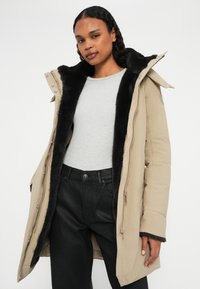Beige parka met zwarte bontvoering, ritsluiting en drukknoopjes; gedragen over een lichtgrijze geribde trui en zwarte broek.