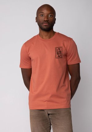 Man draagt een roestkleurig kort mouw T-shirt met een zwart bloemenrechthoekig ontwerp op de borst, staande tegen een effen lichte achtergrond.