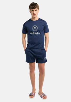 Junger Mann steht mit den Händen in den Taschen, trägt ein marineblaues Carlo Colucci T-Shirt und passende Shorts mit gemusterten Slidesandalen.