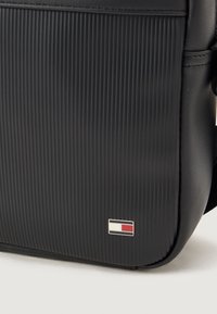 Černá texturovaná taška s vertikálním žebrováním, na boku má malé logo s červenými, bílými a modrými akcenty. Hladké okraje a měkký materiál.