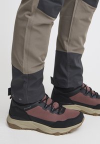 Waterdichte wandelschoenen met een zwart en bordeauxrood gaas bovenwerk, een gestructureerde rubberen zool en een vetersysteem. Tan en zwarte broek met drukknoopsluiting aan de uiteinden van de pijpen.