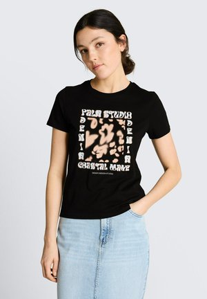 Młoda kobieta z ciemnymi włosami związanymi w niski kok, ubrana w czarny t-shirt z abstrakcyjnymi wzorami i jasnoniebieską dżinsową spódnicę, ręka w kieszeni.