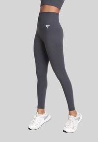 Osoba w ciemnoszarych legginsach z wysokim stanem i białych butach sportowych, stojąca na tle jasnoszarej płyty.