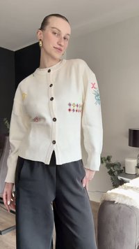 Baltas megztas kardiganas su dygsniuota tekstūra, dekoruotas spalvingais siuvinėjimais ant rankovių ir tamsiomis sagomis. Derinamas su juodomis kelnėmis.