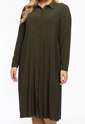 Kobieta ubrana w ciemnooliwkową sukienkę do kolan, z długim rękawem, zapinaną na guziki, z kołnierzykiem na białym tle.