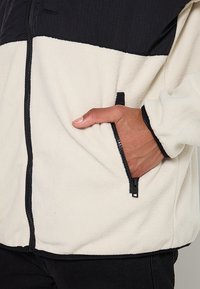 Fleece bunda s krémovým tělem, černými horními panely, zipovou kapsou a texturovanou tkaninou. Obsahuje stojáček a elastické manžety pro pohodlí.
