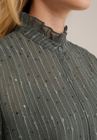 Sötétzöld blúz fodros gallérral, függőleges csíkozással és textúrált részletekkel, kis flitterekkel és gombos záródással.