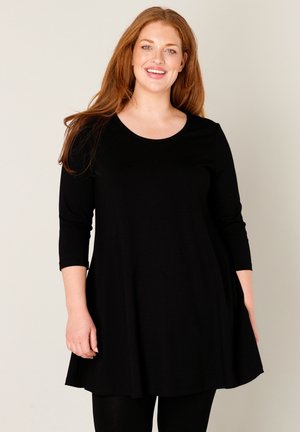 Glunderende vrouw met lang rood haar, gekleed in een loszittende zwarte jurk met driekwartmouwen, tegen een beige achtergrond.