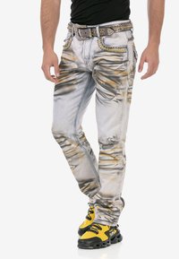 Verblasste Jeans aus hellem Grau mit distressed Effekten, gelben Untertönen, dekorativen Nieten und einem schlanken, geraden Schnitt. Kombiniert mit schwarzen und gelben Schuhen.