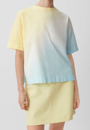 Person iført en kortærmet gradient t-shirt i gul, hvid og lyseblå med et ensfarvet lysgult skørt mod en ensfarvet baggrund.