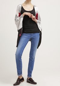 Kimono ligero de frente abierto con patrones florales en negro y rojo en las mangas. Lleva puesto un top negro y lo combina con unos vaqueros azules y zapatos oscuros.