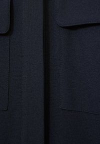Tamsi mėlyna vilnos medžiaga su lygiu paviršiumi, su dviem krūtinės kišenėmis ir vertikaliu centriniu siūlu, einančiu žemyn per priekį.