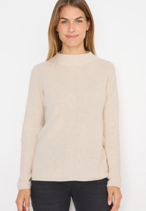 Maglione beige a maglia con collo alto, texture a coste e maniche lunghe, abbinato a jeans neri. Presenta sottili linee verticali.