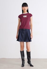 Woman wearing maroon short-sleeve top, navy pleated denim skirt, and black knee-high heeled boots, standing against plain white background.