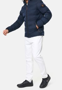 Granatowa kurtka puchowa z zamkiem z przodu i kapturem, z teksturowaną powierzchnią i brązowymi skórzanymi akcentami, połączona z białymi dżinsami i czarnymi sneakersami.