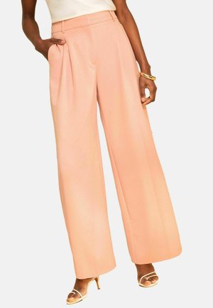 Žena má na sebe broskyňové nohavice s vysokým pásom a širokými nohavicami s záhybmi, v kombinácii s béžovými topánkami na remienky a zlatým náramkom na zápästí.