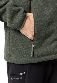 Olivgrüne Fleecejacke mit strukturiertem Finish, ausgestattet mit einer Reißverschlusstasche und elastischen Bündchen. Schwarze Hose mit sichtbarer Reißverschlusstasche.