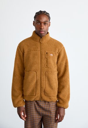 Brown fleece jacket with high collar, zip closure, and two large front pockets. Features a small logo on the chest and textured surface.