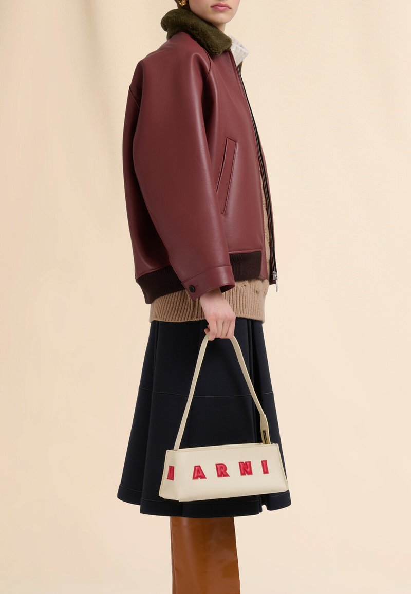 Giubbotto bomber in pelle bordeaux con colletto in pelliccia verde, maglione beige a maglia, gonna nera e borsa crema con scritte rosse.