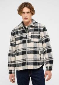 Kurtka polarowa z czarno-białym wzorem w kratę, zapinana na suwak, z dwiema kieszeniami na piersi i kołnierzem. Pokazana z ciemnymi dżinsami.