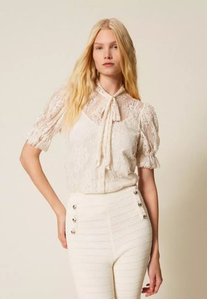 Donna bionda che indossa una camicetta di pizzo trasparente con fiocco al collo e pantaloni bianchi a vita alta con texture e bottoni decorativi.