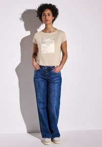 Světlé béžové tričko s čtvercovou grafikou a textem, ve spojení s modrými širokými džíny a šedými teniskami. Hladké textury látky, volný střih.