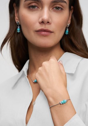 Woman wearing turquoise beaded bracelet and matching earrings, dressed in white blouse, fist raised near face, neutral background.