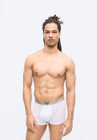 Άντρας με κότσια χωρίς πουκάμισο, φορώντας λευκά εσώρουχα boxer με μπεζ ριγωτή μέση, στέκεται μπροστά σε απλό φωτεινό φόντο.