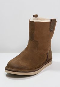 Ruskea nahkainen nilkkuri, jossa on pehmeä sisävuori, pyöreä kärki ja joustava pohja. Siinä on sivupainike ja tikkausyksityiskohtia.